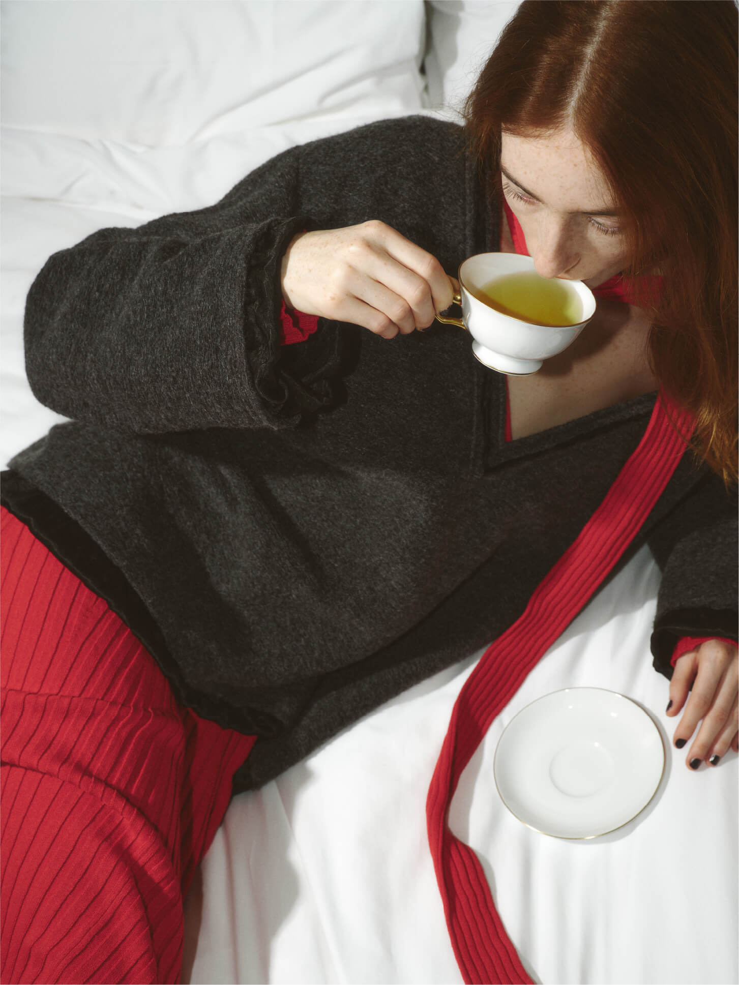 グレイ色のVネックトップスと赤い色の2WAYリブニットワンピースを着用してお茶を飲みながらベットに座ってるモデルイメージ